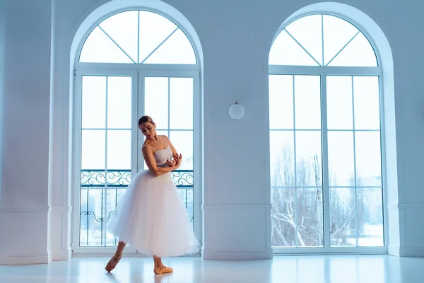 slender ballerina in long white tulle skirt, crouching in bow pose, against backdrop of huge window