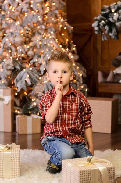 一个金发碧眼的男孩穿着红色格子花衬衫 坐在圣诞树旁 盒子里塞满了礼物 嘴边挂着一个手指 沉默的画像 — 图库照片