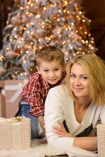 快乐的金发小男孩抱着妈妈躺在地板上 靠近礼品盒 背靠着一棵圣诞树 — 图库照片