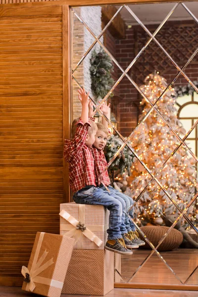 一个穿牛仔裤 穿着格子呢红衬衫的小男孩 双手高举 坐在一堆礼品盒上 镜子映衬着圣诞树 象征着新年的到来 — 图库照片