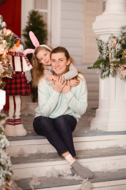 Kulaklı bir çember takan kızı, dekoratif bir kardan adamın ve Noel süslemelerinin yanında, verandada annesine sarılıyor.