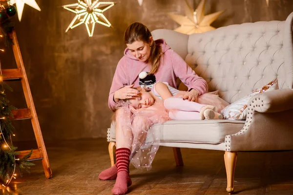 mother strokes the hair of her sleeping daughter on her lap, sitting on the sofa, in a room with a loft interior, against the background of a wall with luminous stars