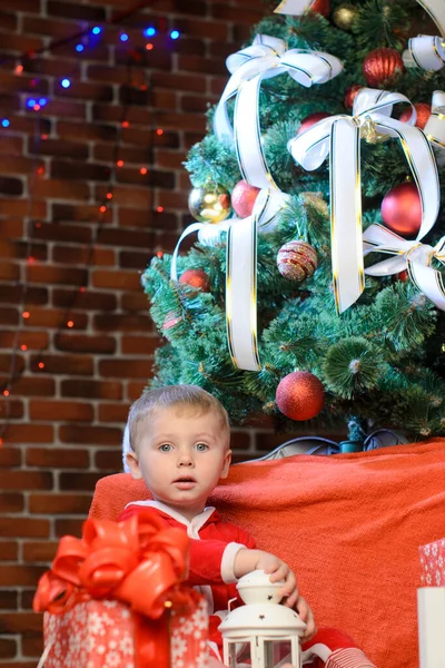 一个身穿圣诞老人服装的小男孩坐在圣诞树下的盒子旁 盒子里装着礼物 靠着砖墙 墙上挂着防波堤 — 图库照片