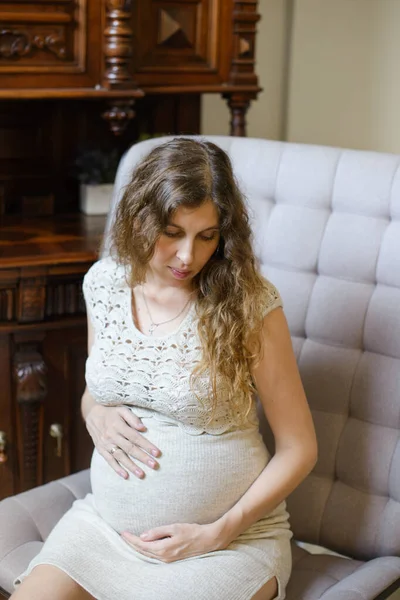 一个穿着针织衣服的孕妇坐在房间里的扶手椅上 看着她的肚子 拥抱着他 — 图库照片