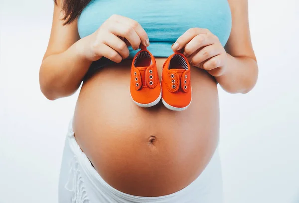 閉じる白い背景に隔離された小さな赤い赤ちゃんのブーツを持つ妊娠中の女性のおなかの写真 新しいライフコンセプト — ストック写真