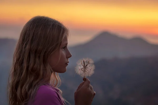 プロフィール穏やかな夕日の光の中で高い山の背景にタンポポの花を持つかわいい女の子の肖像画 平和的な甘い子供 暖かい夜をお楽しみください — ストック写真