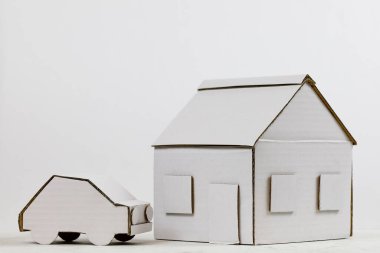 Evin modeli ve yanındaki araba modeli beyaz kartondan yapılmış. Bu iki öğe birlikte mülkü, aile evini veya ipoteği veya diğer gayrimenkul anlamlarını sembolize eder.