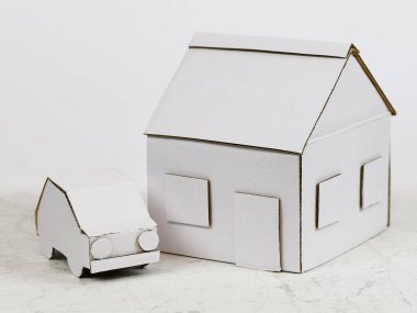 Evin modeli ve yanındaki araba modeli beyaz kartondan yapılmış. Bu iki öğe birlikte mülkü, aile evini veya ipoteği veya diğer gayrimenkul anlamlarını sembolize eder..