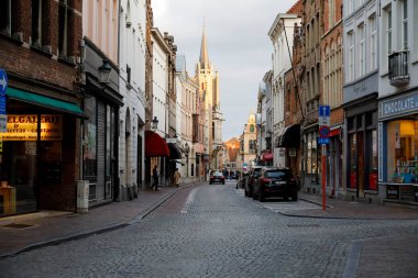 Bruges, Belçika - 14 Eylül 2022: Dar, her iki tarafında tarihi evler bulunan asfalt şehir sokağı. Güneş ışınları kulenin bir kısmını aydınlatıyor. Alacakaranlık yaklaşıyor.