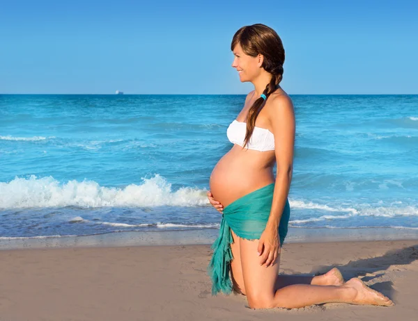Красивая беременная женщина на коленях на голубом пляже — стоковое фото