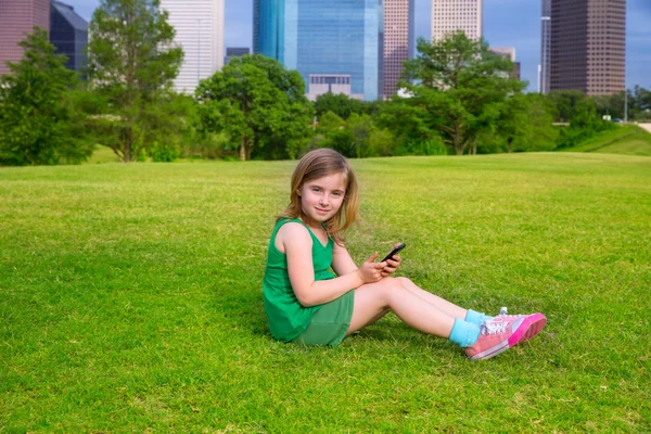 Rubia niña jugando con el teléfono inteligente sentado en el césped del parque en c —  Fotos de Stock