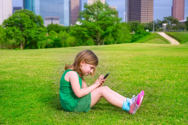 Blond kid girl playing with smartphone sitting on park lawn at c — Stock Photo, Image