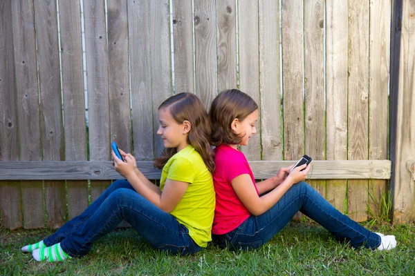 Twin sister girls playing smartphone sitting on backyard lawn — Stock Photo, Image