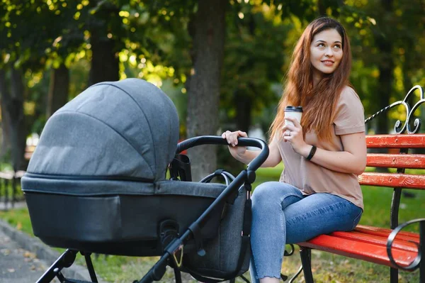 Şık, genç anne parkta bir bebek arabasıyla bankta oturuyor.