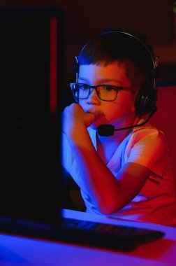 Child online broadcasts computer game, boy streams in headphones on rgb lighting background clipart