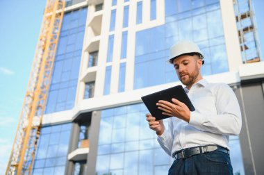 Vinçlerin yanındaki mühendis. Konsept - büyük inşaat projesi. Mimar inşaat sürecini yönetir. Ellerinde çizimler ve tablet.