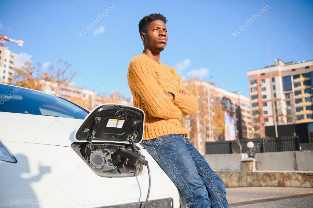 Close up of african american man connecting charging cable to electric car. Young male standing near his modern auto with leather suitcase in hand.