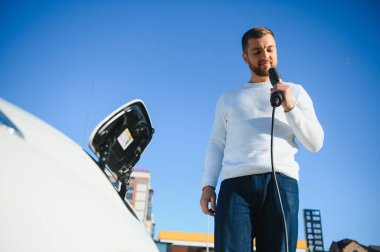 Genç yakışıklı adam elektrik şarj istasyonunda kablo tutuyor. Yeni arabasının yanında duruyor..