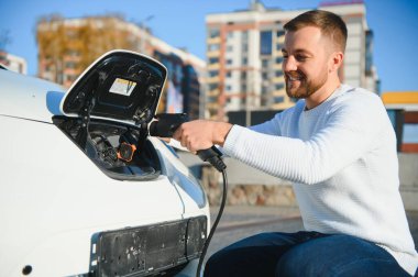 Genç yakışıklı adam elektrik şarj istasyonunda kablo tutuyor. Yeni arabasının yanında duruyor..