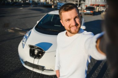 Genç, yakışıklı Avrupalı bir adamın portresi, akıllı telefonuna selfie çekerken elektrikli arabasına yaslanıyor, alışveriş merkezinden yeni ürünler aldıktan sonra şehrin elektrik santralinin pilini şarj ediyor.