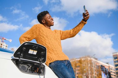 Elektrikli arabalar, EV konsepti, çevre dostu yakıt. Genç gülümseyen zenci adamın portresi modern lüks elektrikli arabasını şarj ediyor.
