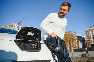 Genç yakışıklı adam elektrik şarj istasyonunda kablo tutuyor. Yeni arabasının yanında duruyor..