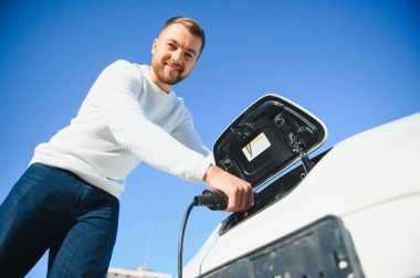 Genç yakışıklı adam elektrik şarj istasyonunda kablo tutuyor. Yeni arabasının yanında duruyor..