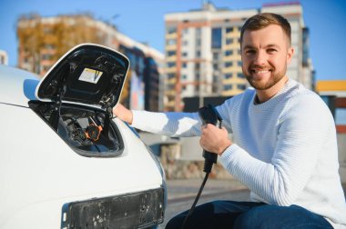 Genç yakışıklı adam elektrik şarj istasyonunda kablo tutuyor. Yeni arabasının yanında duruyor..