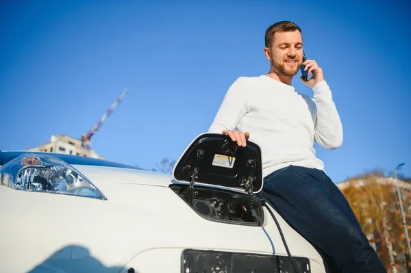 男は車のフードの上に座った 彼の車は充電ステーションで充電している 男はスマートフォンの画面を見て微笑む — ストック写真