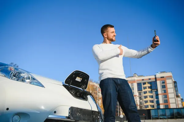 若いハンサムな男が充電ケーブルを持っている彼の新しい車の近くに立って充電ステーションポイント — ストック写真