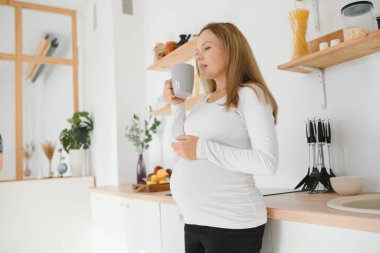 Güzel, sıradan giyinmiş esmer bir kadının bebek beklerken, modern şık bir mutfakta sabah çayı içerken, gülümserken fotoğrafı. Beklenti, beklenti, çocuk doğurma ve annelik kavramı