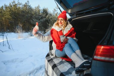Genç bir kadın arabanın bagajında oturmuş sıcak çay içiyor ve kış karlı bir günde selfie çekiyor..