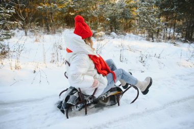 Güzel, genç bir kadın sıcak bir kış elbisesi giyiyor, kızağın üstünde oturuyor ve tepeden aşağı kayıyor, karlı bir kış gününde eğleniyor.