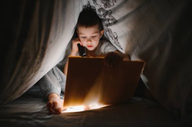 Çocuk yatakta kitap okuyor. Çocuklar geceleri okur. Yatak odasında peri masalı kitapları olan küçük bir çocuk. Küçük çocuklar için eğitim. Akşamları masal anlatırsın. Karanlık odada, battaniyenin altında, lambalı sevimli bir çocuk..