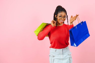 Büyük indirim. Alışveriş çantalı, güneş gözlüklü, pembe stüdyo duvarının üstünden kameraya bakan heyecanlı afro kız.