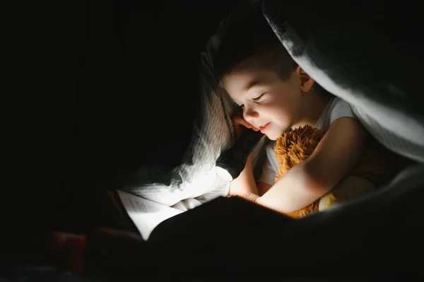 A boy of 5-6 years old is reading a book in the evening in the dark under a blanket with a toy bear