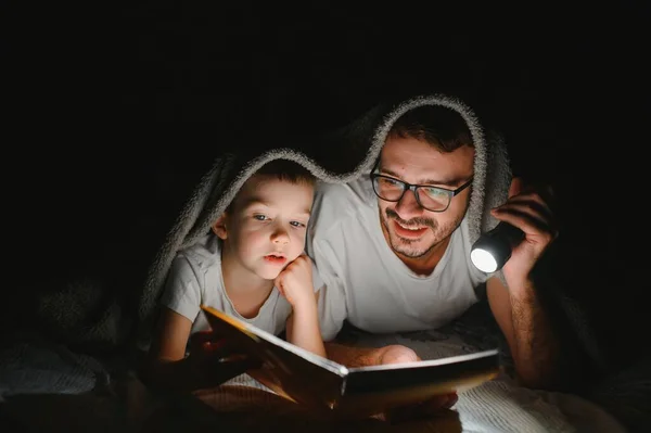 Happy family reading bedtime story under blanket in evening. Father and son spend time together. Father\'s Day.