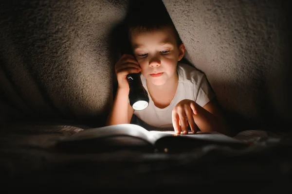 Child reading book in bed. Kids read at night. Little boy with fairy tale books in bedroom . Education for young children. Bedtime story in the evening. Cute kid under blanket in dark room with lamp.