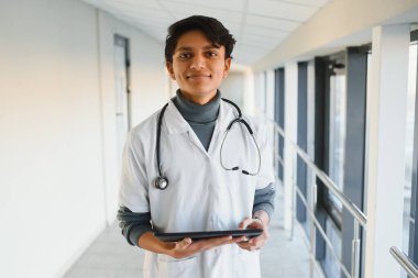 Beyaz önlüklü, kendine güvenen genç Arap erkek doktor portresi, elinde bir panoyla modern hastane binasının arka planında duruyor.