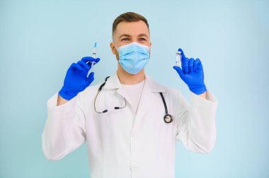 Koronavirüse karşı aşı. COVID-19 aşıları test ediliyor. Tıbbi maskeli bir erkek doktor mavi arka planda COVID-19 aşısı olan bir şırınga dolduruyor. Doktorun elleri şırıngayı yakın çekimde tutuyor..
