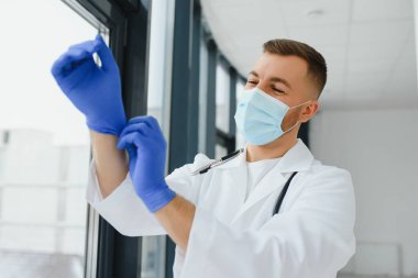 Beyaz üniformalı, gözlüklü, boynunda steteskop olan ve hasta muayenesinden önce lastik eldiven takan beyaz erkek doktor..