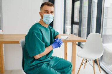 Koruyucu maskeli genç adam içeride otururken kameraya bakıyor.