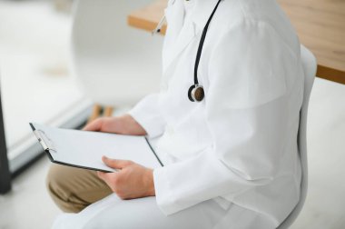 Yakın çekim, beyaz tıbbi paltolu kırpılmış bir adam. Doktor hastanın kartına bir şey yazıyor. Işık fotoğrafı.