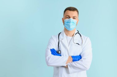 Tıbbi maske takan genç bir erkek doktor ve boynunda steteskop var. Mavi arka planda izole edilmiş.