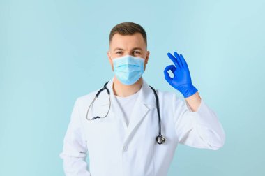Tıbbi maske takan genç bir erkek doktor ve boynunda steteskop var. Mavi arka planda izole edilmiş.
