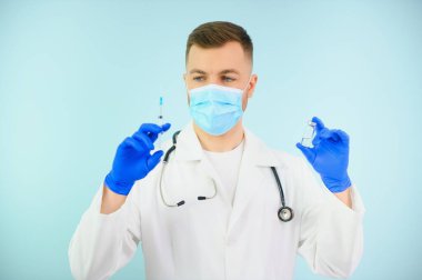 Koronavirüse karşı aşı. COVID-19 aşıları test ediliyor. Tıbbi maskeli bir erkek doktor mavi arka planda COVID-19 aşısı olan bir şırınga dolduruyor. Doktorun elleri şırıngayı yakın çekimde tutuyor..