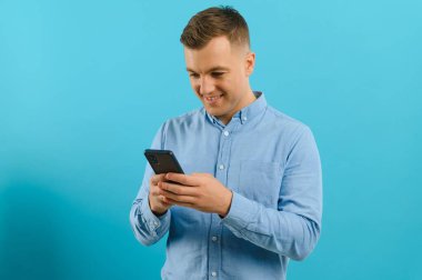 Cep telefonunu elinde tutan, mesajlaşan mutlu yakışıklı adamın fotoğrafı. Mavi arka planda poz veren büyük dişli genç adam..