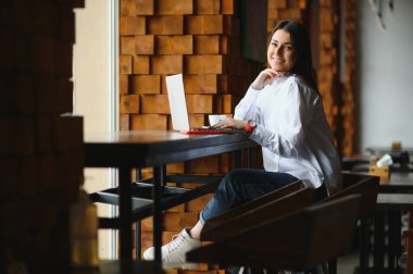 Şirin kız, gülümseyen ve şık bir kafede dizüstü bilgisayarla çalışan.