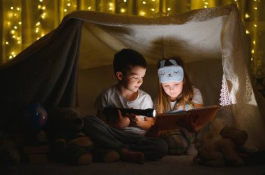 Mutlu aile çocukları evdeki çadırda el feneriyle kitap okuyor.