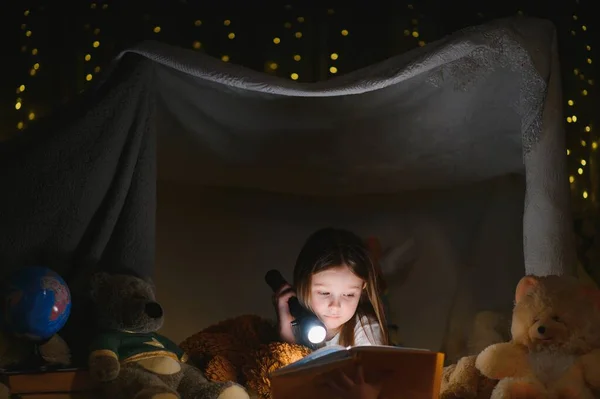 少女はテントの中で懐中電灯の本を読んでいる 幸せな女の子が家で遊んでいる 面白い素敵な子供が部屋で楽しんでいます — ストック写真